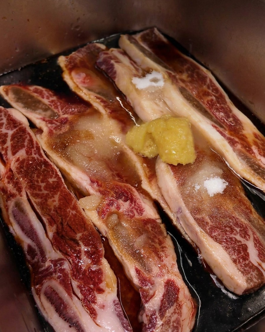 A close-up shot of a mound of pale yellow minced garlic resting on top of raw, marbled beef short ribs soaking in a dark soy-based marinade.