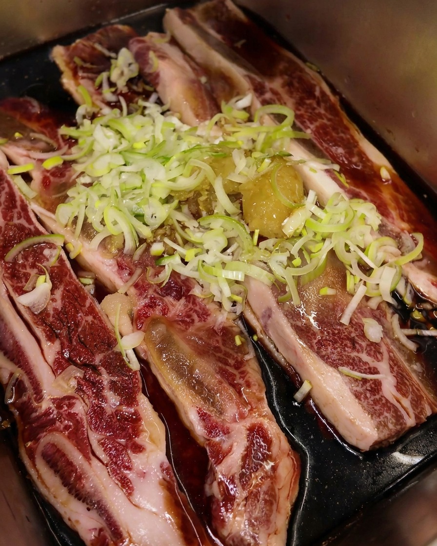 Toasted sesame oil being poured over a tray of beef ribs topped with green onions and garlic, creating a shimmering layer on the dark marinade.