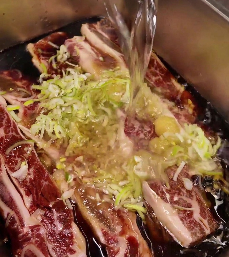 A stream of clear water being poured into a stainless steel tray containing beef ribs topped with minced garlic and sliced green onions.