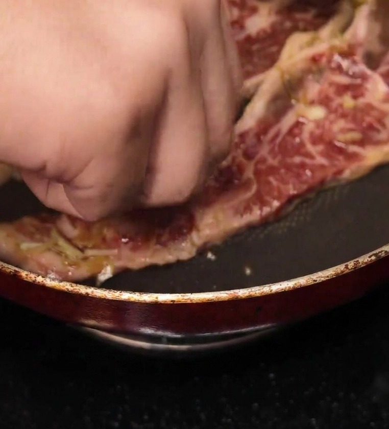 A hand placing a strip of marinated, raw bone-in short rib into a hot non-stick frying pan.
