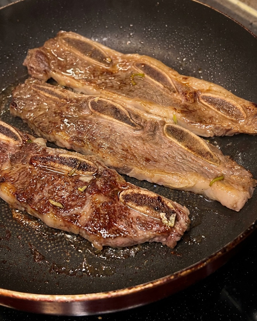 Three strips of LA short ribs sizzling in a black pan, with one being flipped to reveal a dark, caramelized crust.