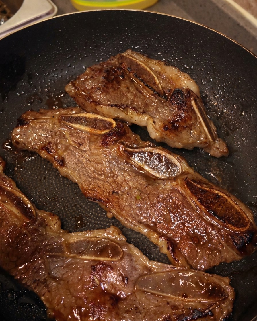 Kitchen shears cutting through a strip of browned beef short ribs inside a frying pan, separating them into small bone-in sections.