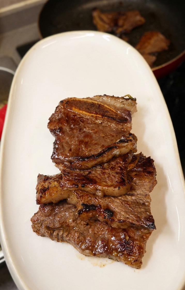 A stack of succulent, dark brown grilled LA short ribs being piled onto a long, oval white ceramic plate.