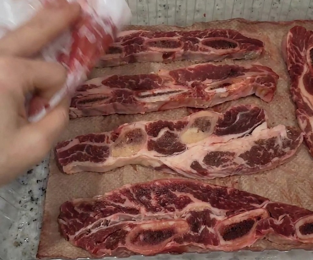 A persons hand using a white paper towel to blot dry raw beef short rib strips laid out in a clear plastic tray.