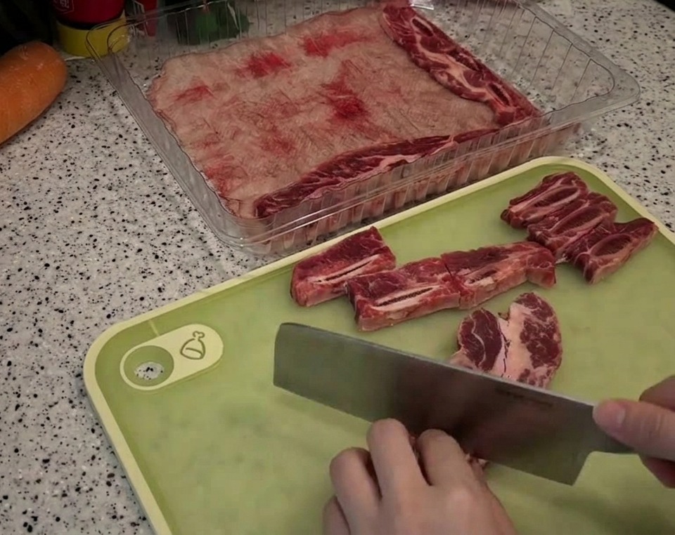 A person using a large cleaver to cut raw, marbled beef short ribs into small rectangular chunks on a light green cutting board.