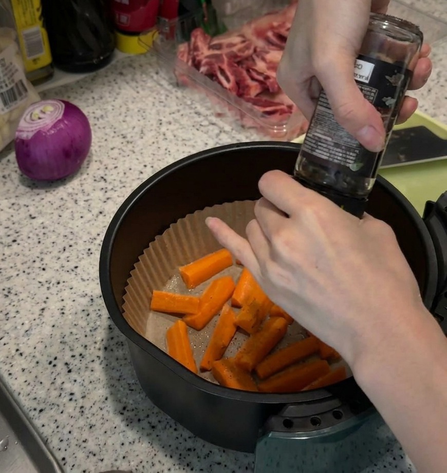 Hands grinding black pepper from a glass mill over a pile of raw orange carrot sticks resting in a paper-lined air fryer basket.