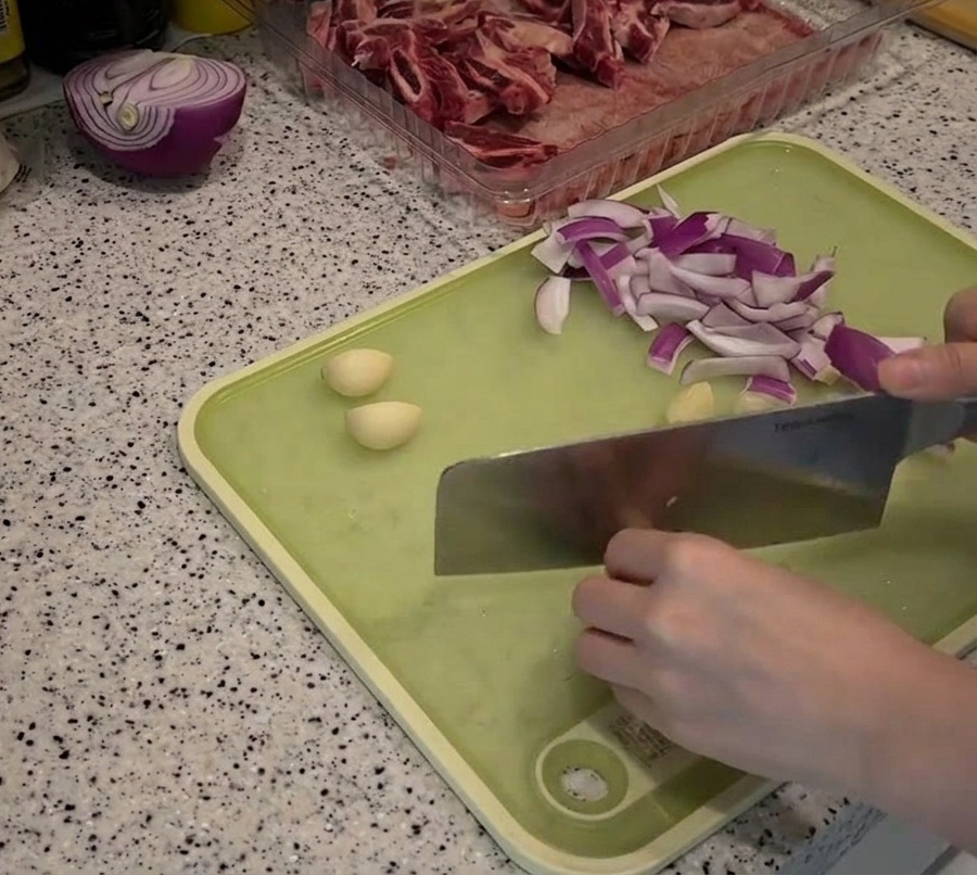 Sliced red onion strips and whole peeled garlic cloves spread across a green cutting board being prepped with a knife.