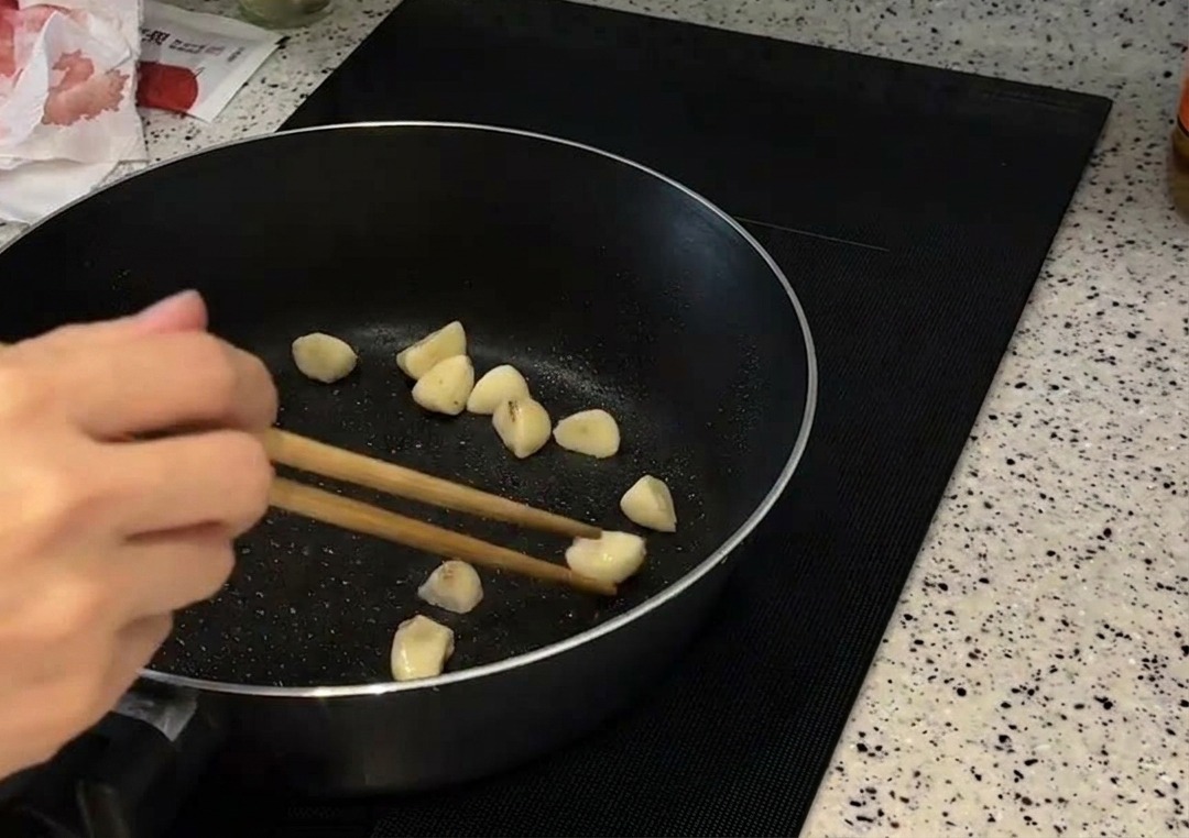 A handful of whole garlic cloves being stirred with wooden chopsticks in a dark frying pan as they turn golden brown.