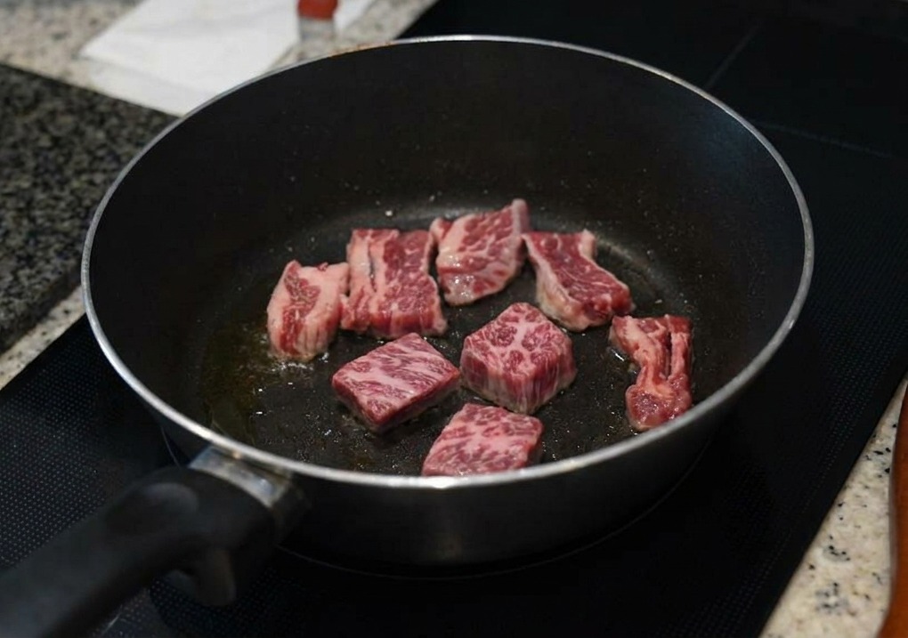 Several pieces of marbled beef short ribs searing in a dark pan, with the cooked edges turning brown and opaque.