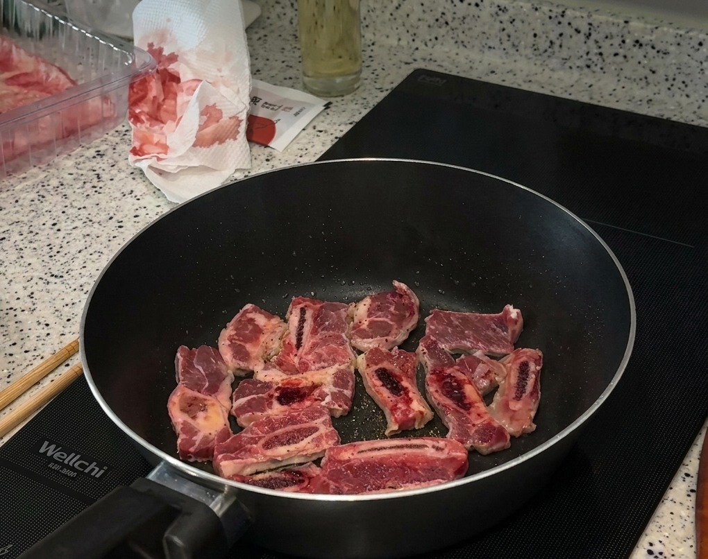 Beef short rib pieces searing in a black pan on an induction cooktop, with visible black pepper and salt seasoning on the meat.
