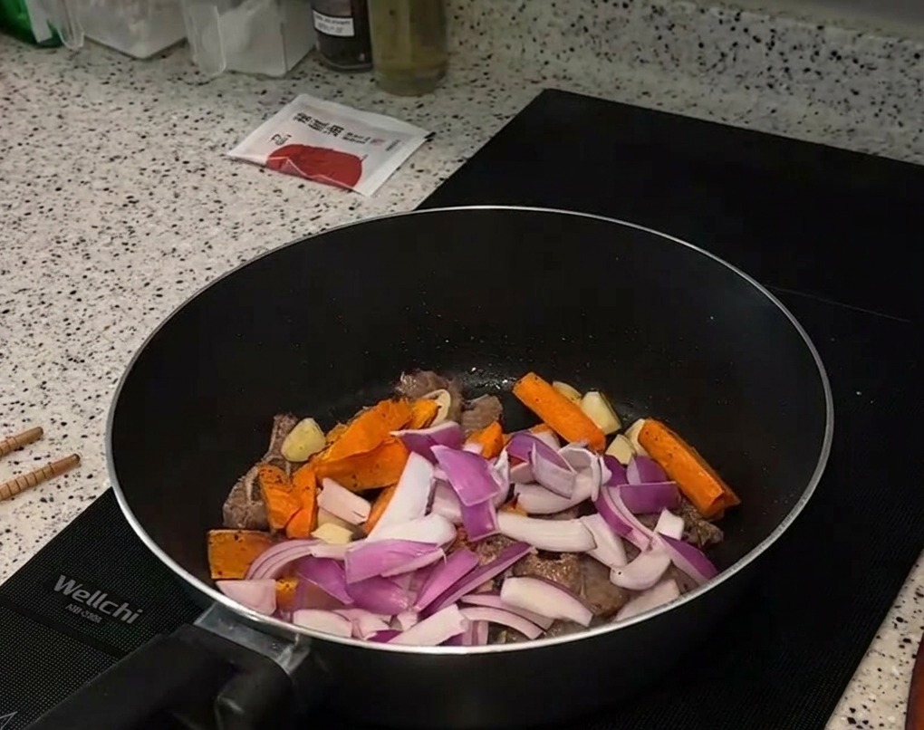 Slices of red onion and roasted orange carrot sticks being added on top of seared beef pieces in a frying pan.