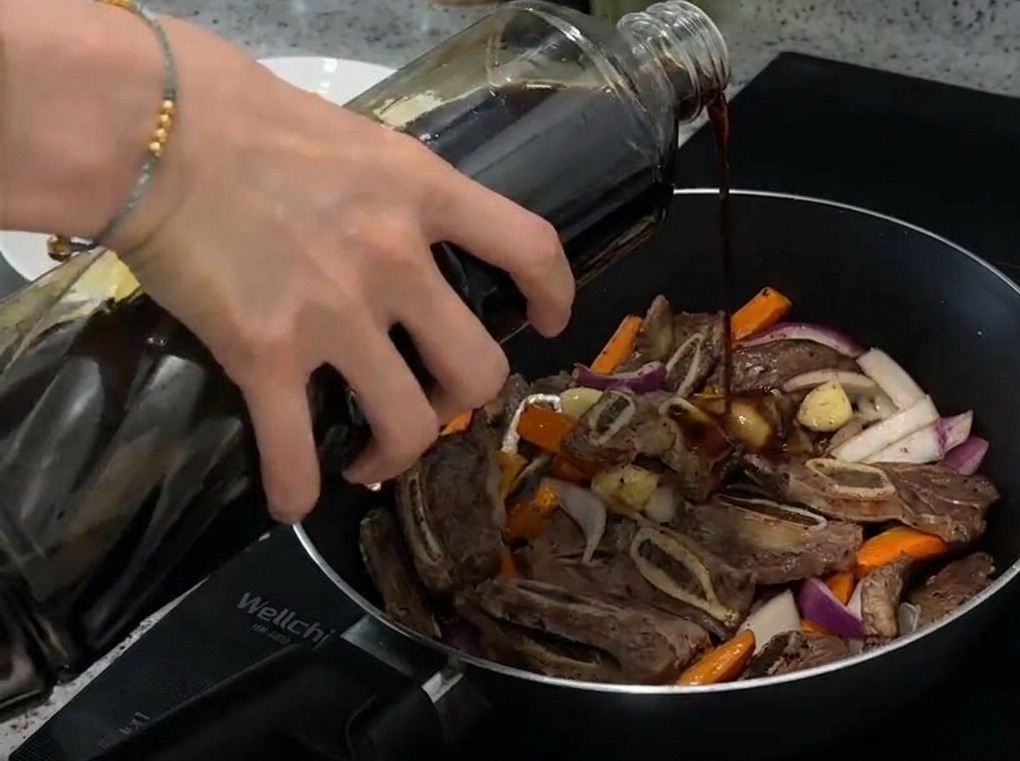 A close-up shot of light soy sauce being poured from a glass bottle into a frying pan containing browned beef ribs, carrots, and sliced red onions.