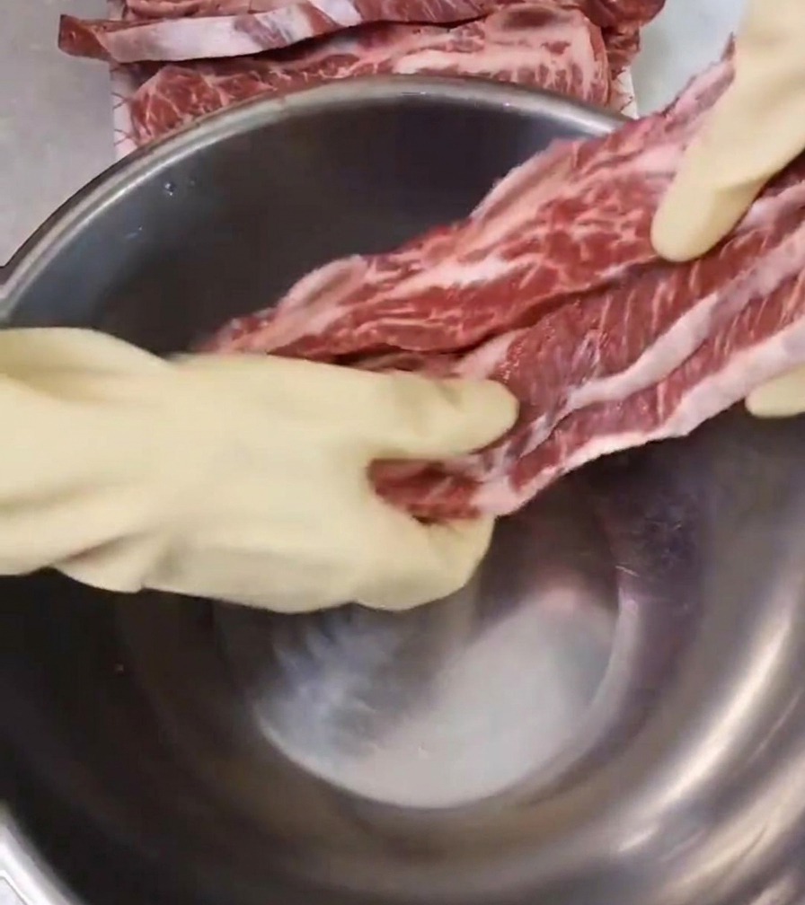 A person wearing white gloves is shown placing raw, marbled beef short ribs into a deep stainless steel mixing bowl on a clean countertop.