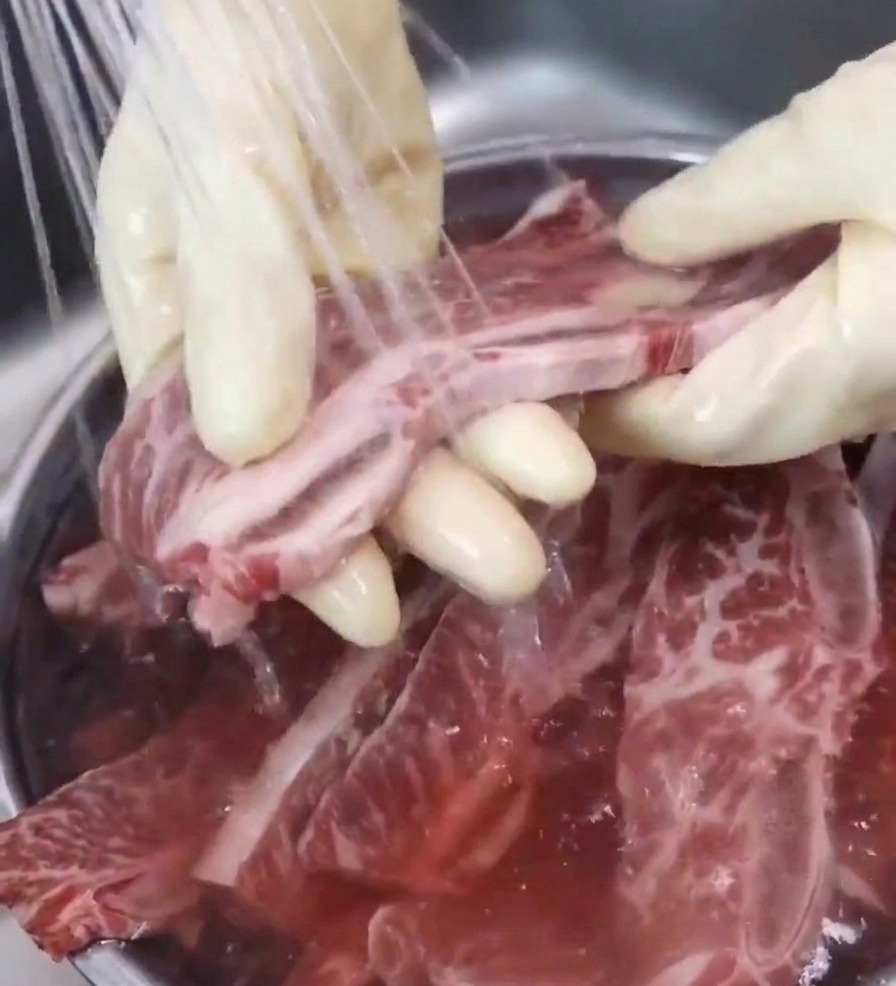 A gloved hand holds a beef short rib under a steady stream of water from a kitchen faucet, rinsing it over a bowl to remove impurities.