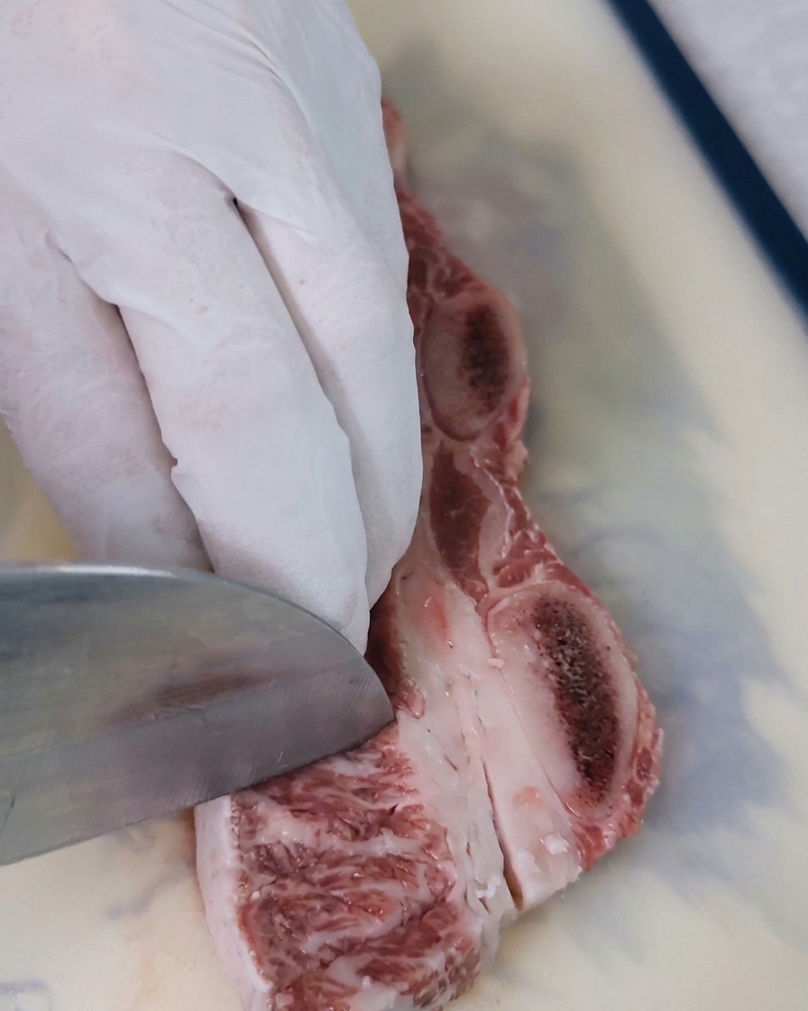 A close-up shot of a knife blade making diagonal cuts across the red flesh of a raw beef rib to create a textured surface for marinating.
