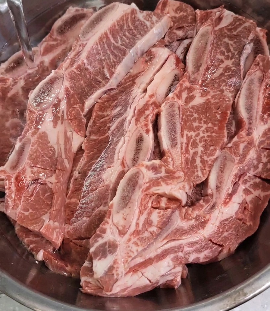 A stream of clear water is poured into a stainless steel bowl, filling it up to cover a large quantity of raw, scored beef short ribs.