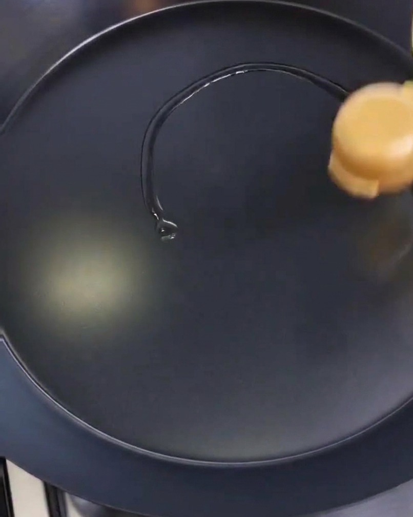 A person is spreading a thin film of oil across the surface of a circular black frying pan using a folded paper towel.