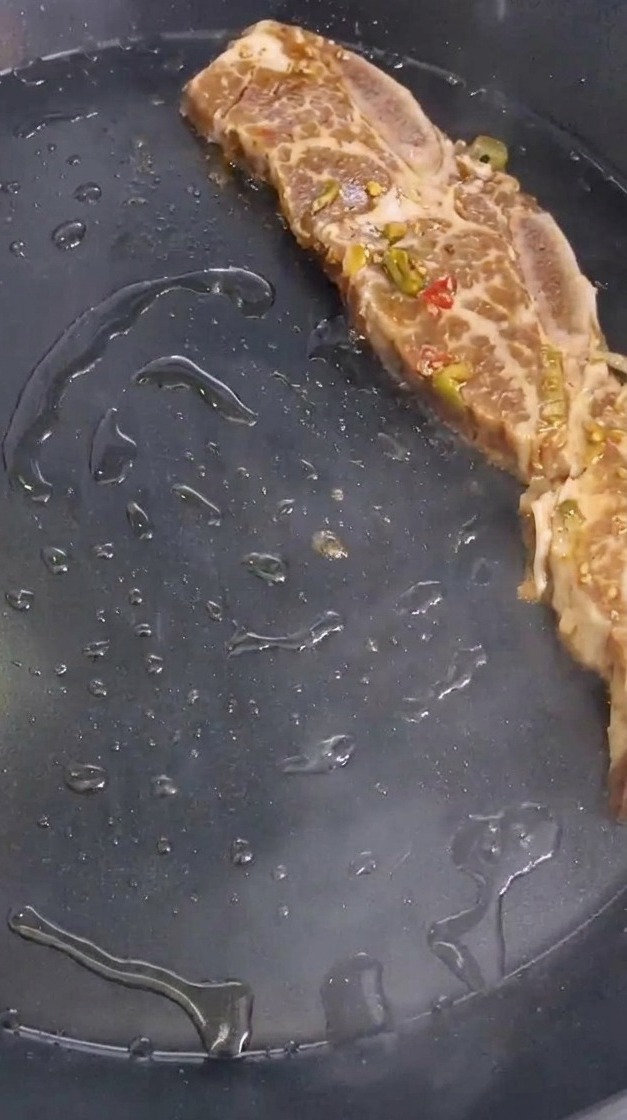 A single marinated beef short rib seasoned with bits of peppers and onions being placed onto a hot, oiled black pan.