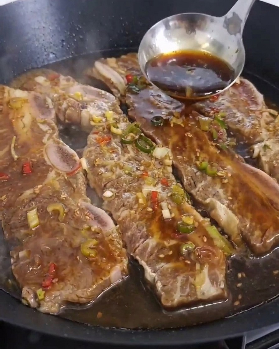 A stainless steel ladle pouring dark brown marinade over several pieces of beef short ribs cooking in a large pan.