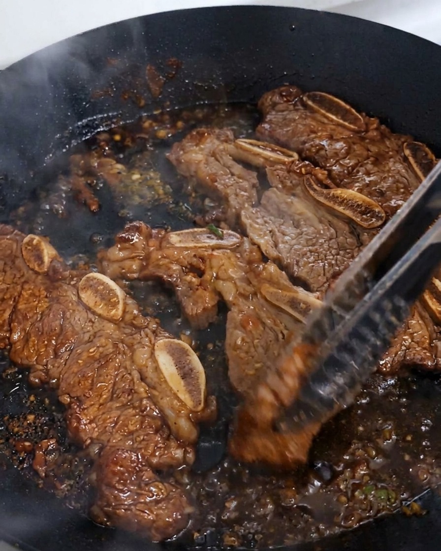 Beef short ribs being turned with tongs in a pan where the dark sauce is bubbling and reducing into a thick, glossy glaze.