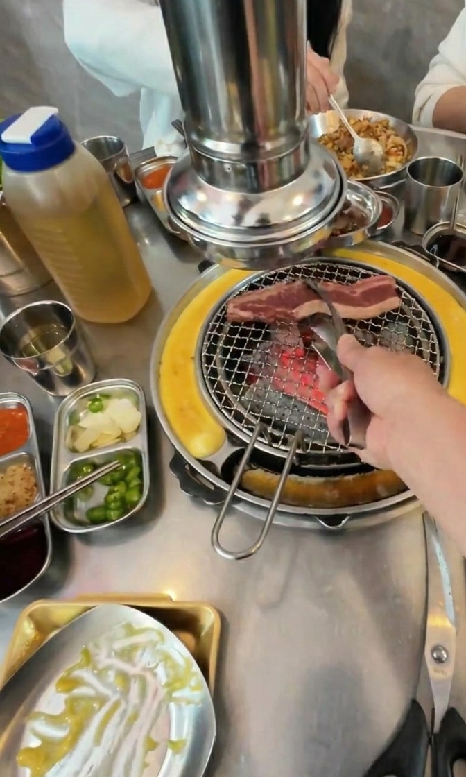 A hand using metal tongs to lay raw, fatty beef strips onto a hot charcoal grill grate surrounded by side dishes.
