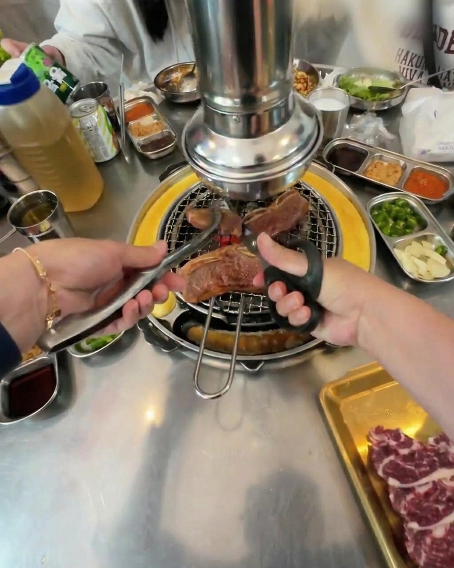 A person using scissors and tongs to cut large pieces of partially cooked beef on a circular grill.