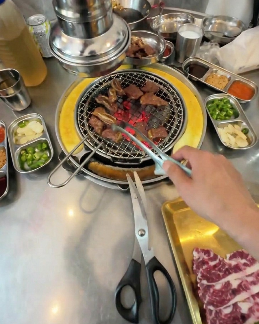 Small cubes of fully grilled beef being tossed with tongs on a charcoal grill grate.