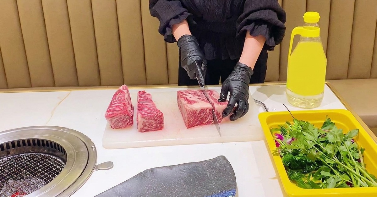 A person in black gloves using a chefs knife to divide a large, marbled raw beef short rib into uniform rectangular sections on a white board.