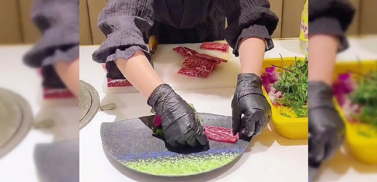 Hands wearing black gloves carefully layering thin, marbled beef slices into an overlapping fan shape on a dark, curved plate.