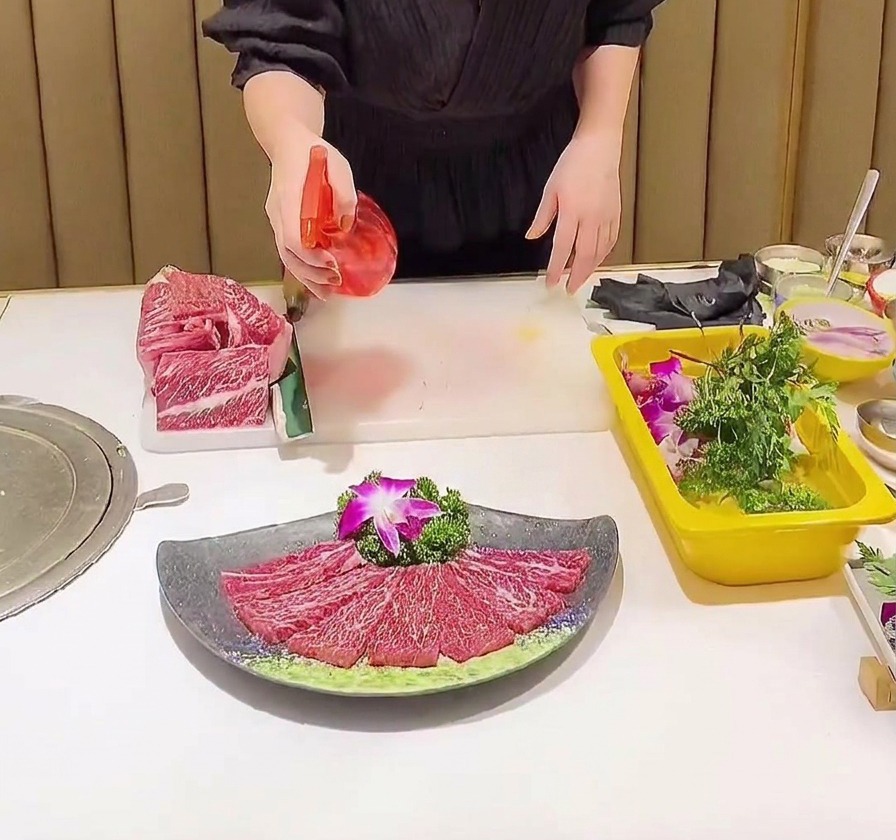 A red spray bottle misting a clear liquid seasoning over a plate of thinly sliced and salted marbled beef.