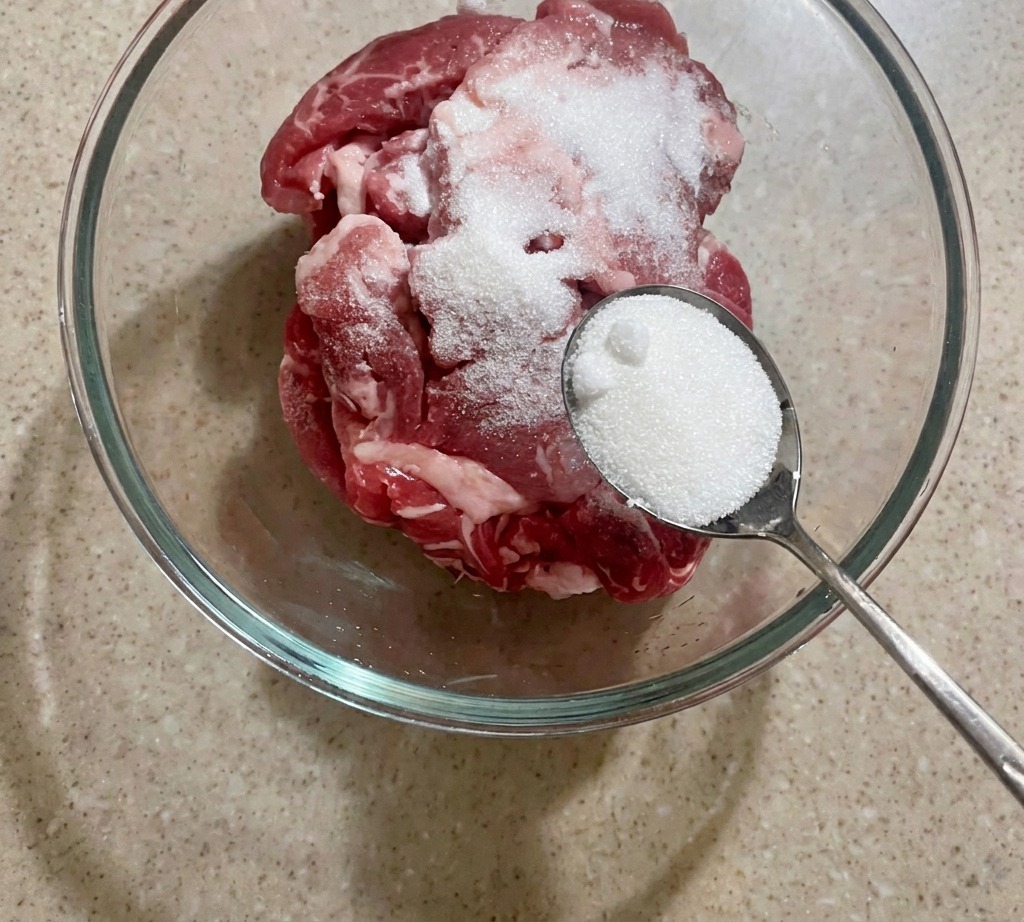 A bowl of raw sliced pork being seasoned with a spoonful of white sugar.