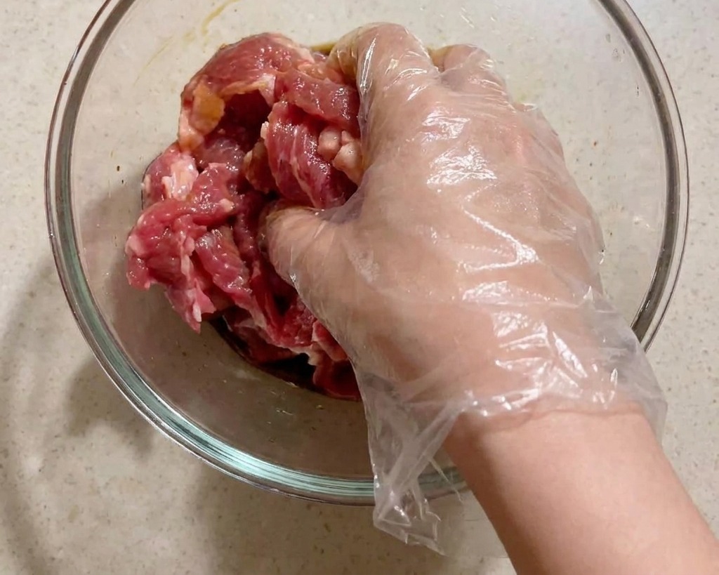 A hand in a clear plastic glove mixing raw pork slices with liquid seasonings in a glass bowl.
