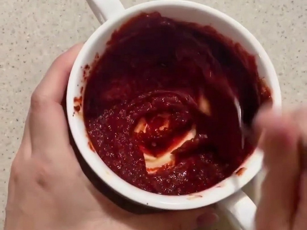 A person mixing a thick, deep red gochujang-based sauce in a small white bowl with a spoon.