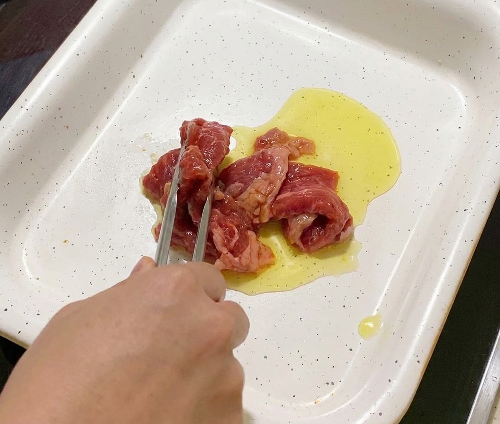 Tongs placing slices of marinated pork into a white pan with shimmering oil.