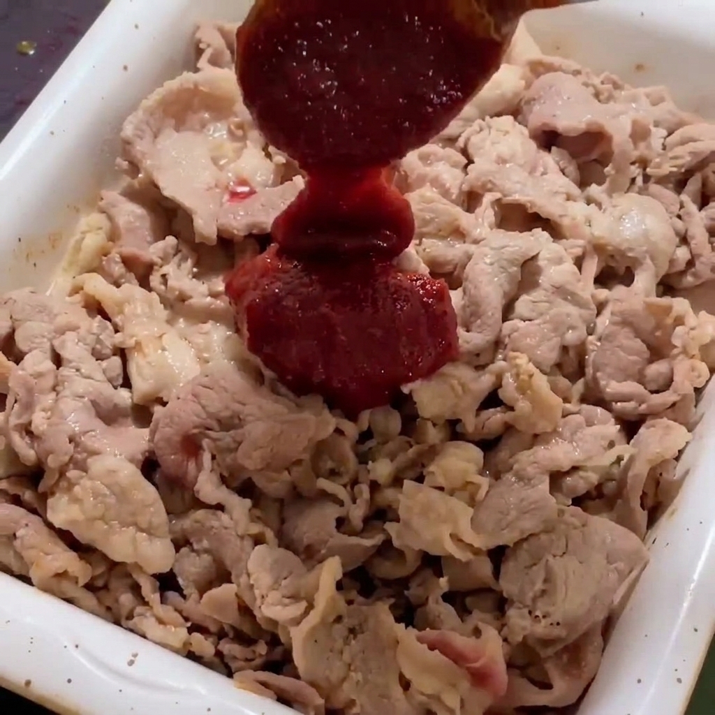 Thick red spicy sauce being added to browned pork slices in a white frying pan.