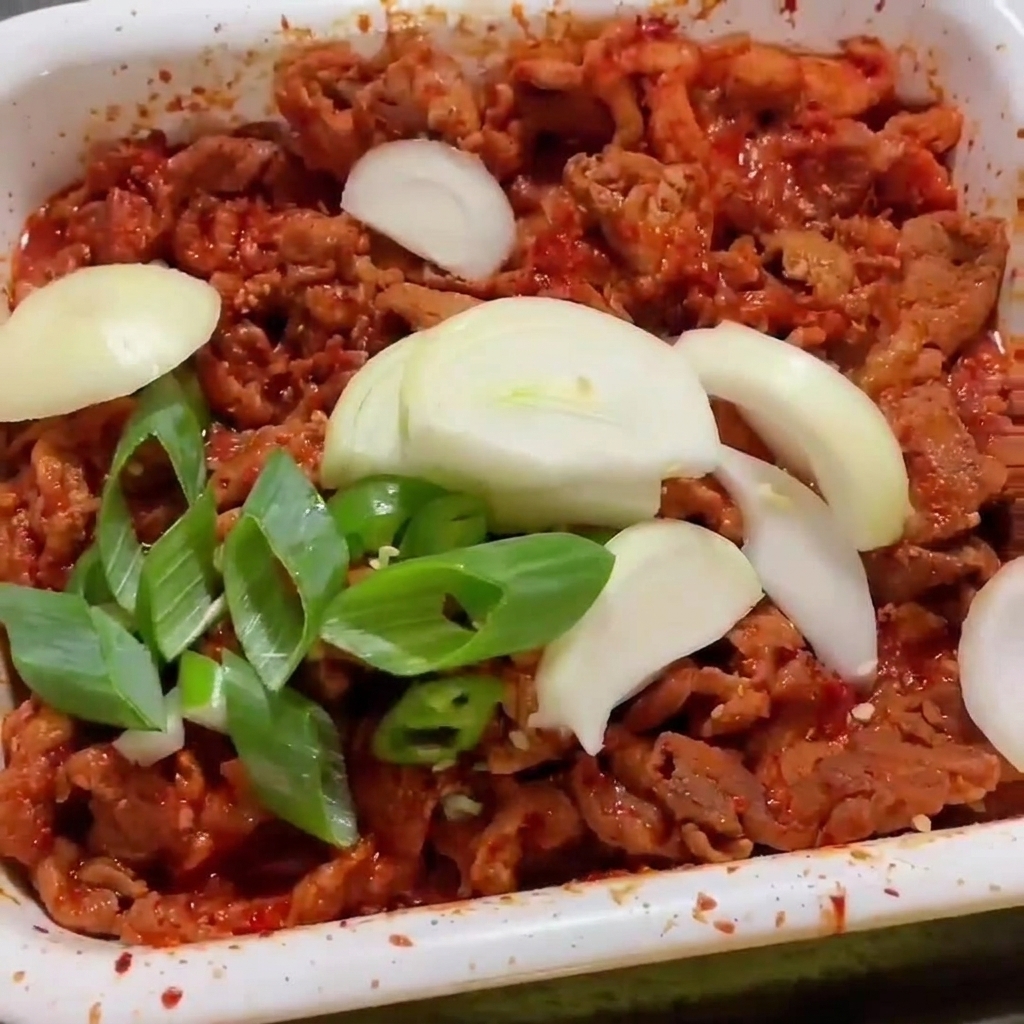Freshly sliced white onions and green onions layered over red, spicy marinated pork strips sizzling in a white pan.