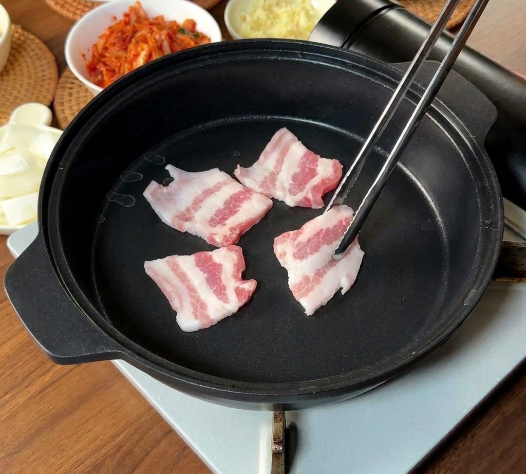 Thin strips of raw, marbled pork belly being placed into a black pan using long metal chopsticks.