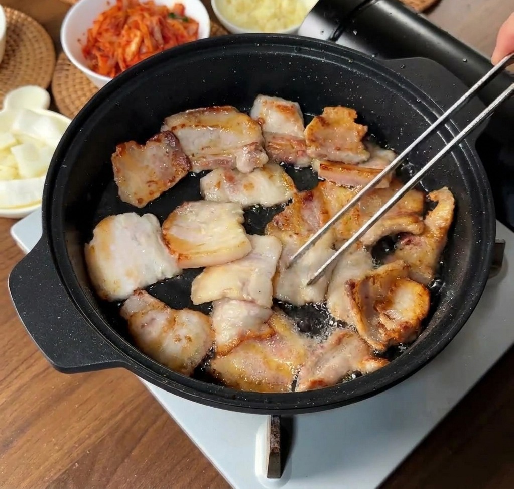 Sliced pork belly pieces browning in their own rendered fat inside a black pan, with some pieces being flipped by tongs.