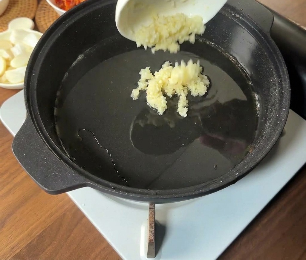 Minced garlic being poured from a small white bowl into the hot, oily center of a black pan.