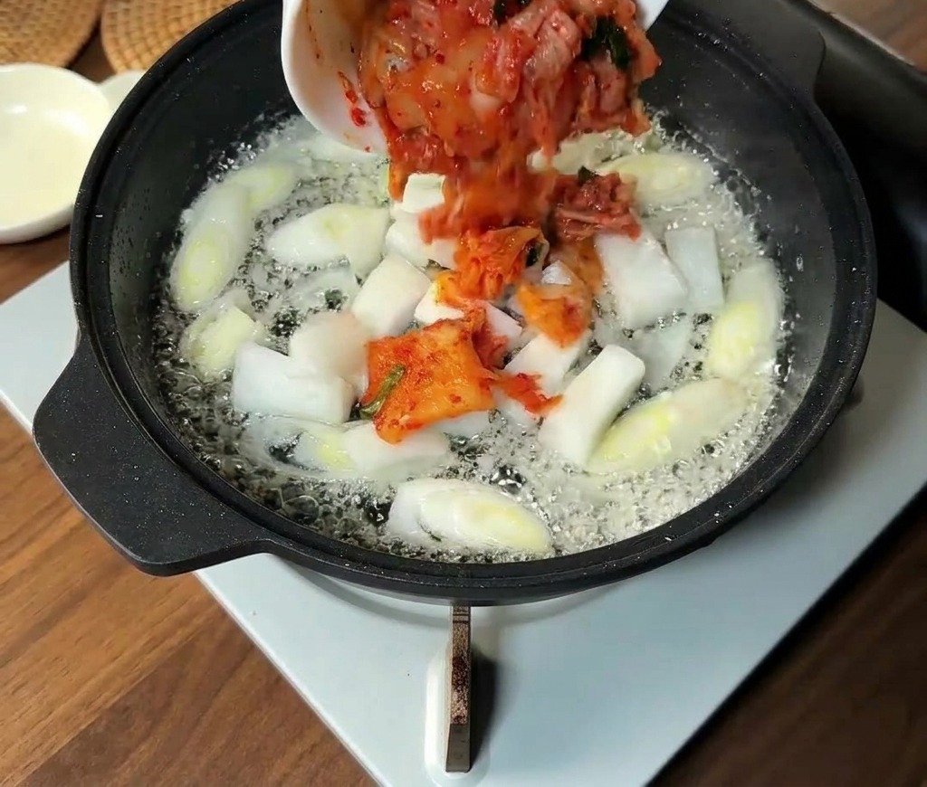 Chopped Korean kimchi and white onion slices being added to a pan containing sautéed scallions.