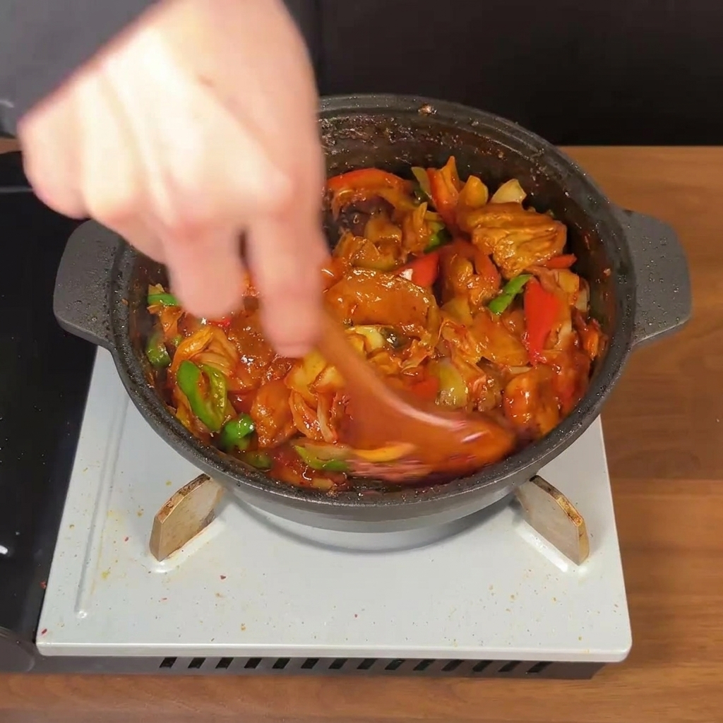 A wooden spoon vigorously stirring a pan of Korean spicy pork belly, kimchi, and peppers in a thick red sauce.