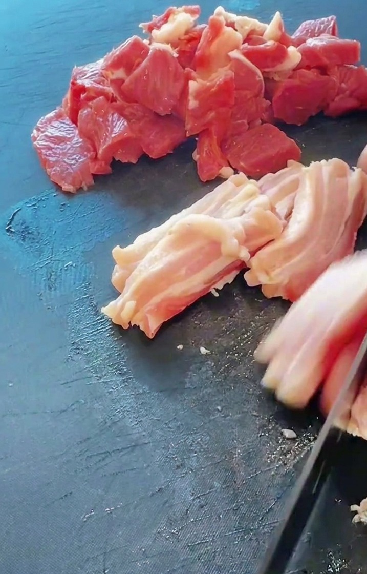 A close-up shot of a knife cutting through fresh pork belly on a dark cutting board with pre-cut meat nearby.