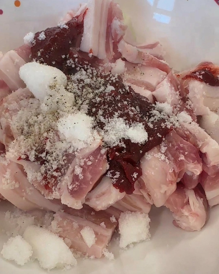 Raw pork strips in a bowl topped with red chili paste, white granulated sugar, and ground black pepper.