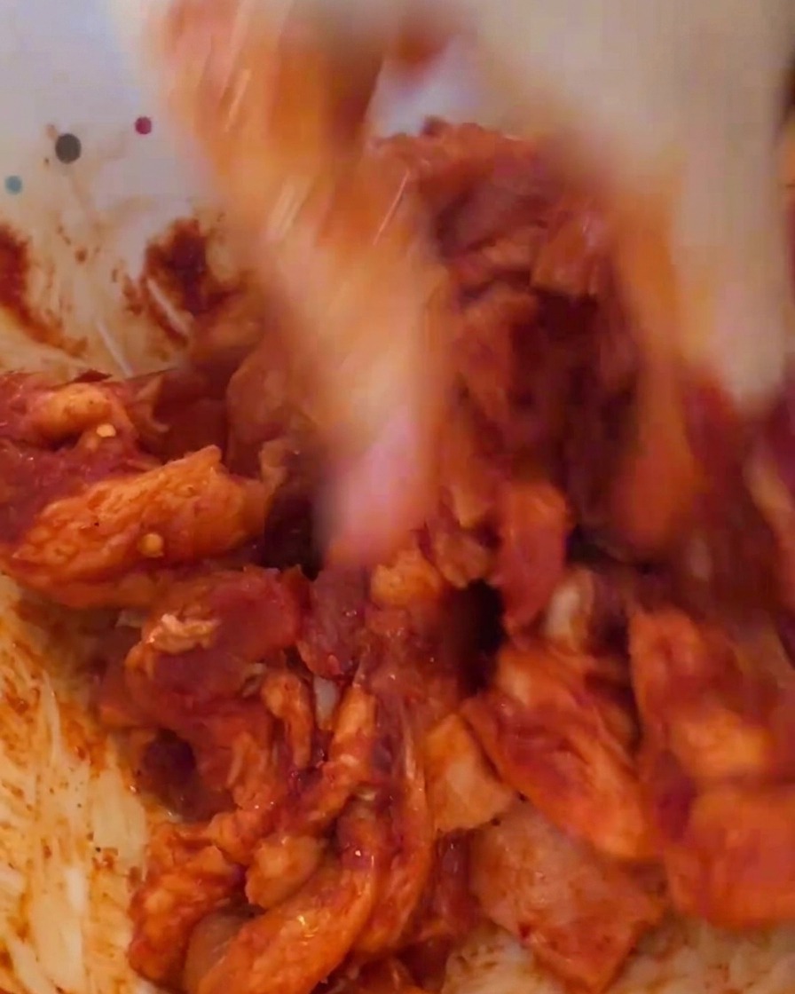 A hand in a plastic glove mixing red marinated meat in a white bowl, showing a vibrant red texture.