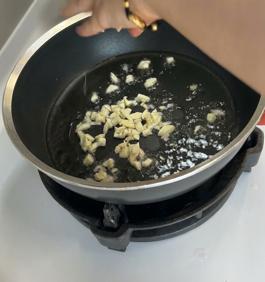 Small bits of chopped garlic frying in oil at the bottom of a dark non-stick pan.