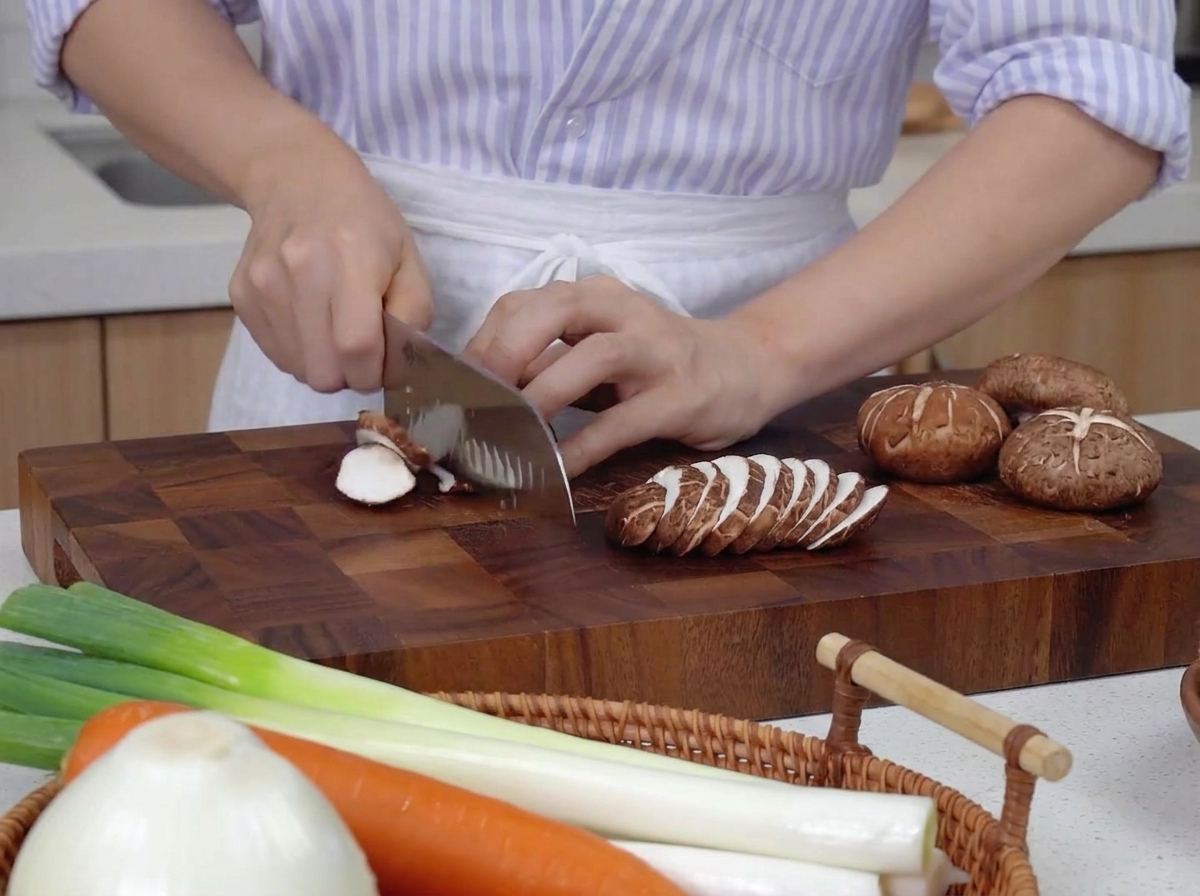 Hands using a chefs knife to thinly slice brown shiitake mushrooms on a dark wood cutting board next to whole carrots and green onions.