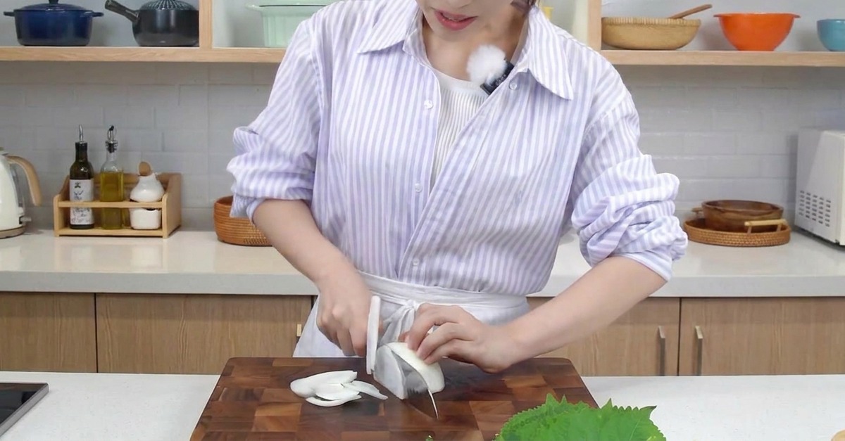 A person using a kitchen knife to slice a white onion into thin strips on a dark wooden cutting board.