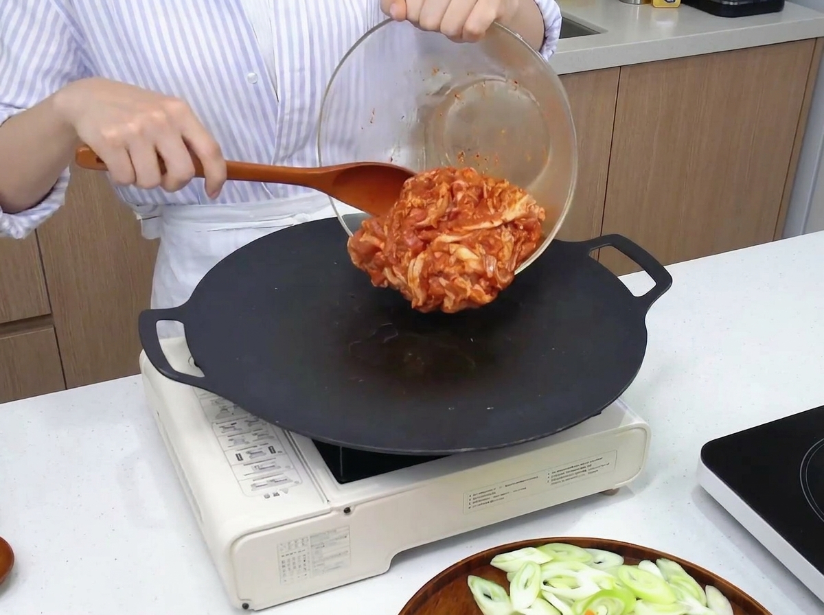Marinated red pork belly being transferred from a glass bowl onto a hot, black circular griddle using a wooden spatula.