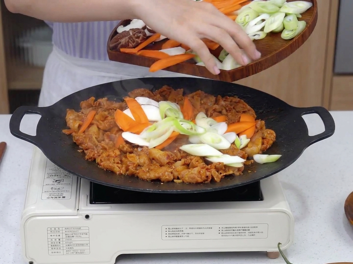 A variety of sliced vegetables including onions, carrots, and mushrooms being dropped onto sizzling spicy pork in a large black pan.