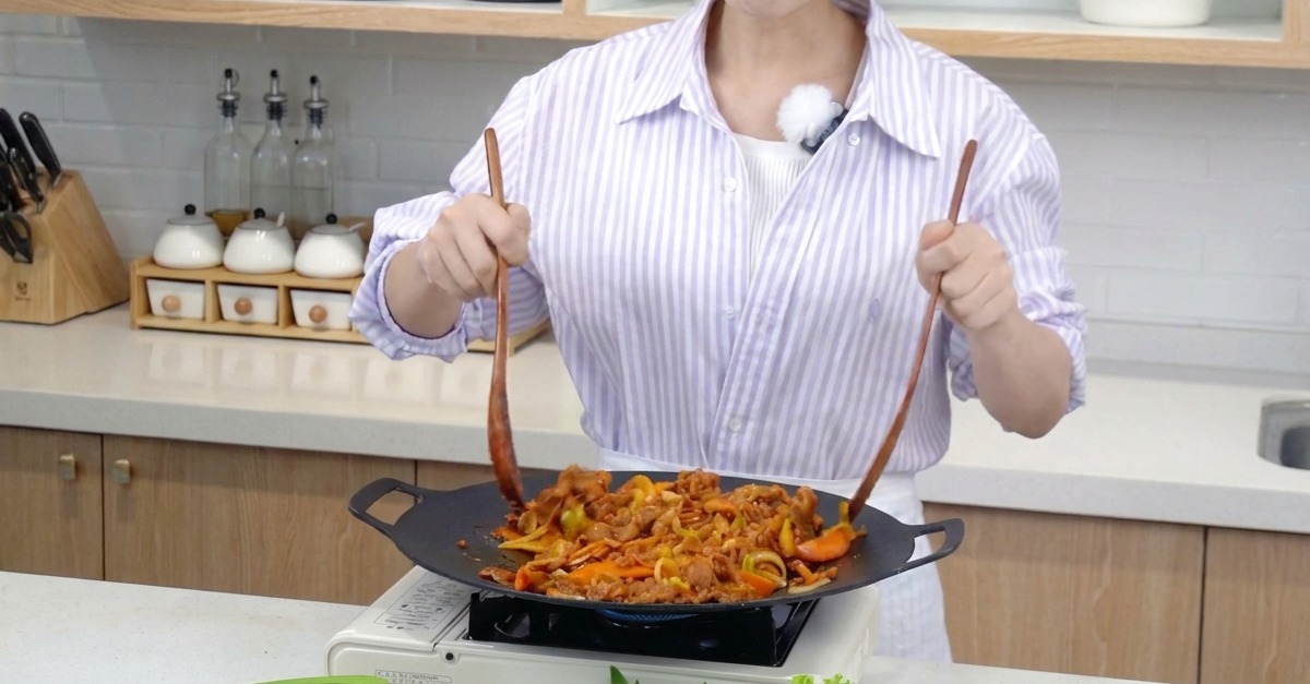 Marinated pork and colorful vegetable strips being tossed in a wide flat pan using two long wooden spatulas.